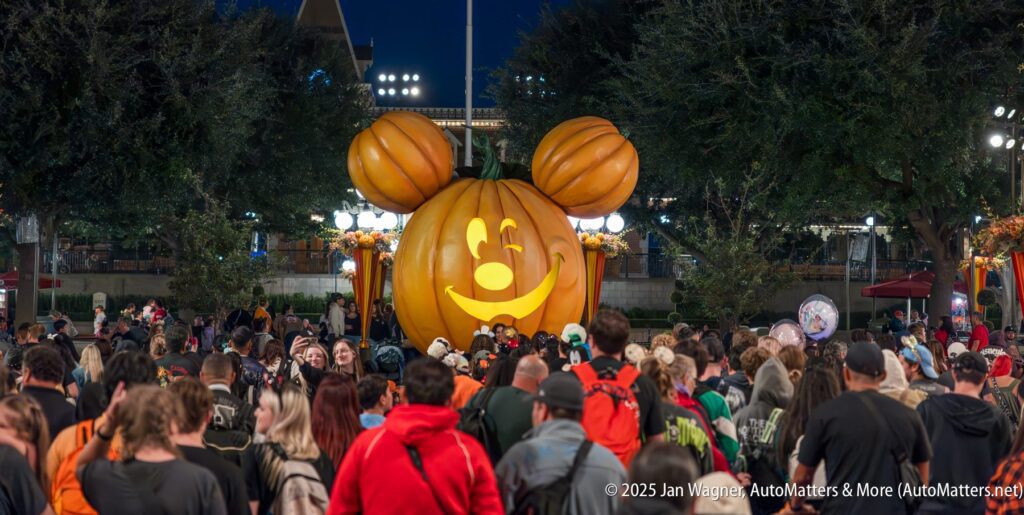 A large Mickey Mouse pumpkin decoration is displayed at night, surrounded by a crowd of people at an outdoor Halloween event.