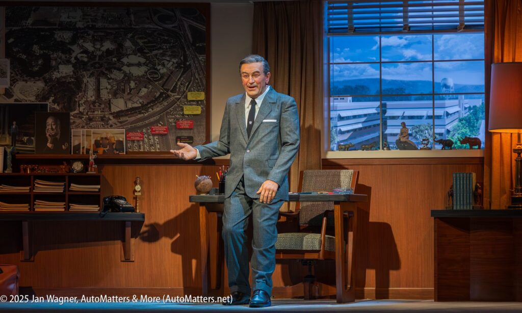 A man in a gray suit stands in an office set with wood furniture, a large map on the wall, and a window showing a building and greenery outside.