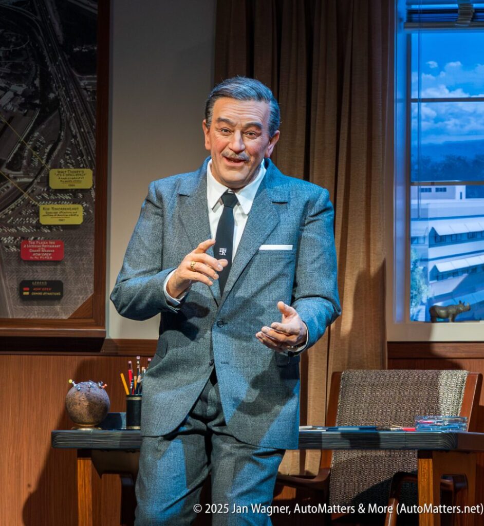 A wax figure of a man in a gray suit stands in an office setting, gesturing with one hand, with a desk and window behind him.