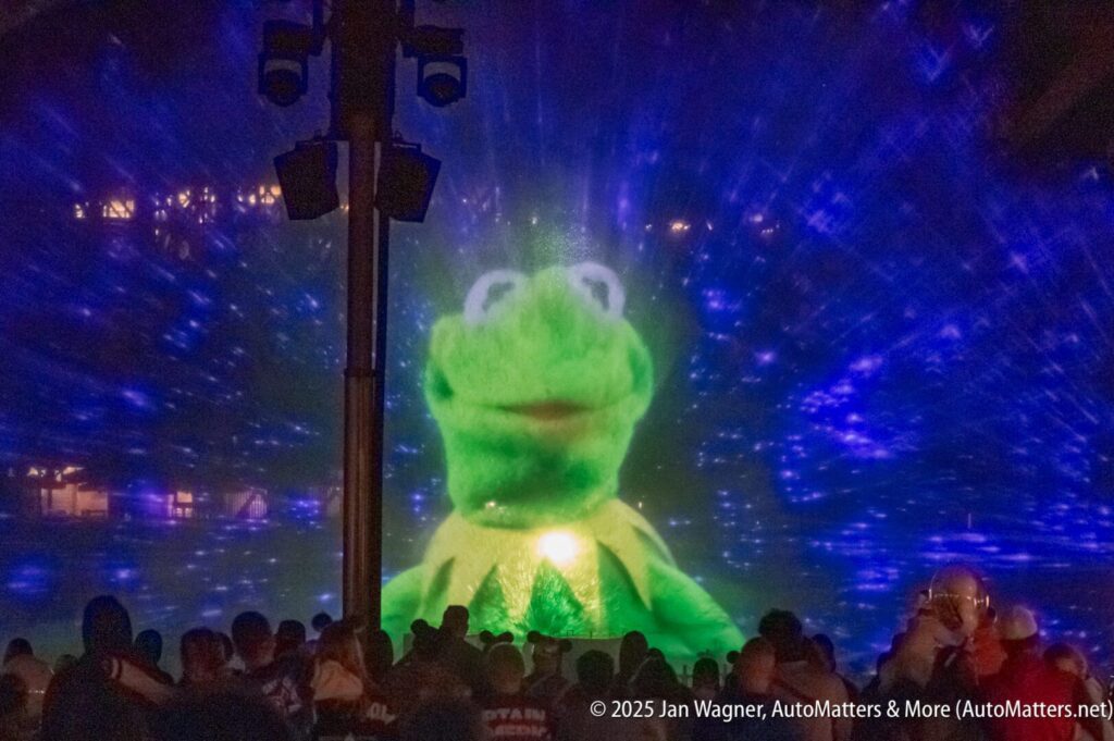 A projected image of Kermit the Frog appears on a water screen at night, with a crowd watching in the foreground and blue lights in the background.