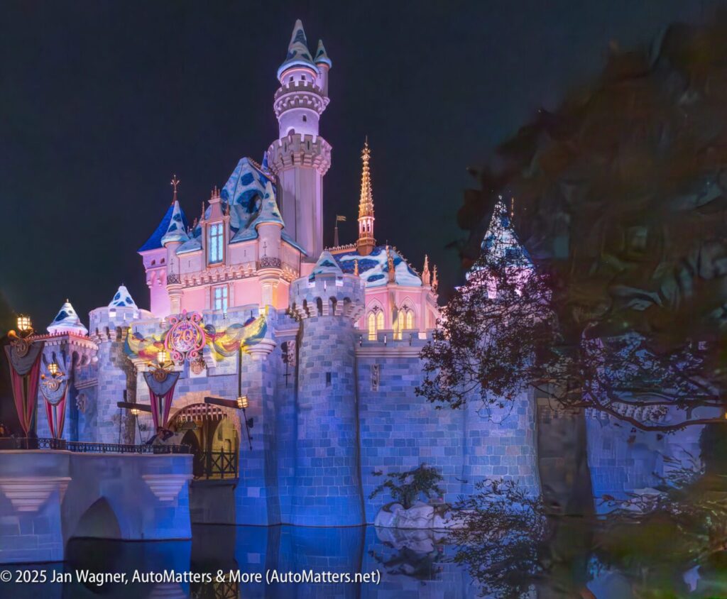 Sleeping Beauty Castle at Disneyland is brightly illuminated at night, with decorative lights and castle flags visible; trees frame the right side of the image.