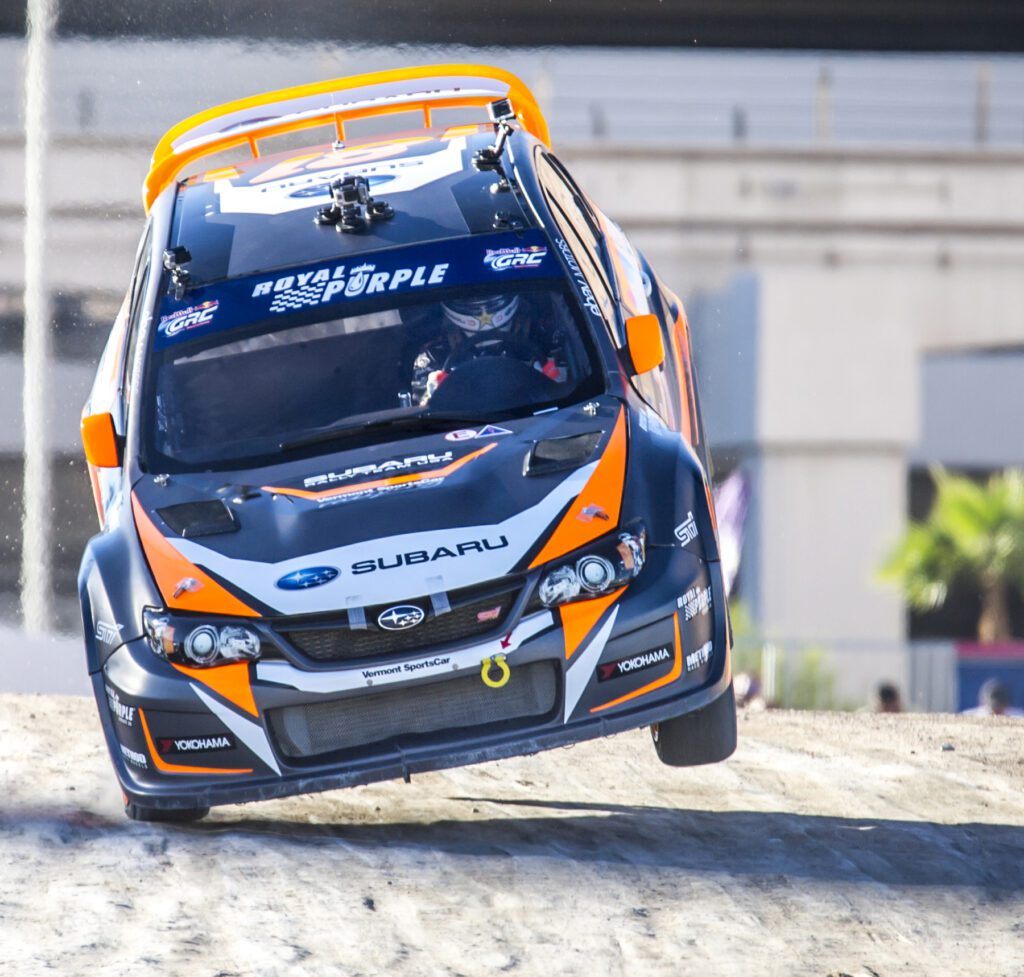 A rally car with Subaru branding drives on two wheels during a race, with the driver visible inside and a blurred background indicating motion.