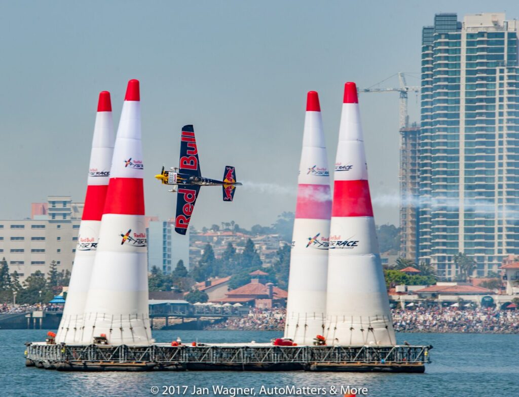 A Red Bull stunt plane flies between inflatable pylons on a water course during an air race, with city buildings and a crowd in the background.