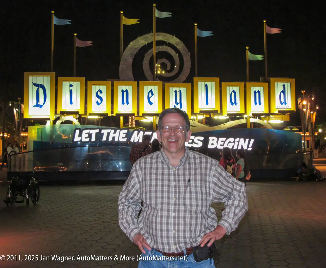 A man in a plaid shirt stands smiling in front of a brightly lit Disneyland sign at night. The sign reads, “LET THE MEMORIES BEGIN!”.