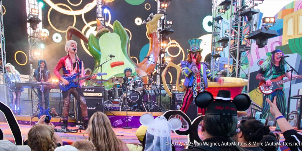A rock band dressed in colorful costumes performs on an outdoor stage with whimsical decorations, while an audience watches, some wearing Mickey Mouse ears.