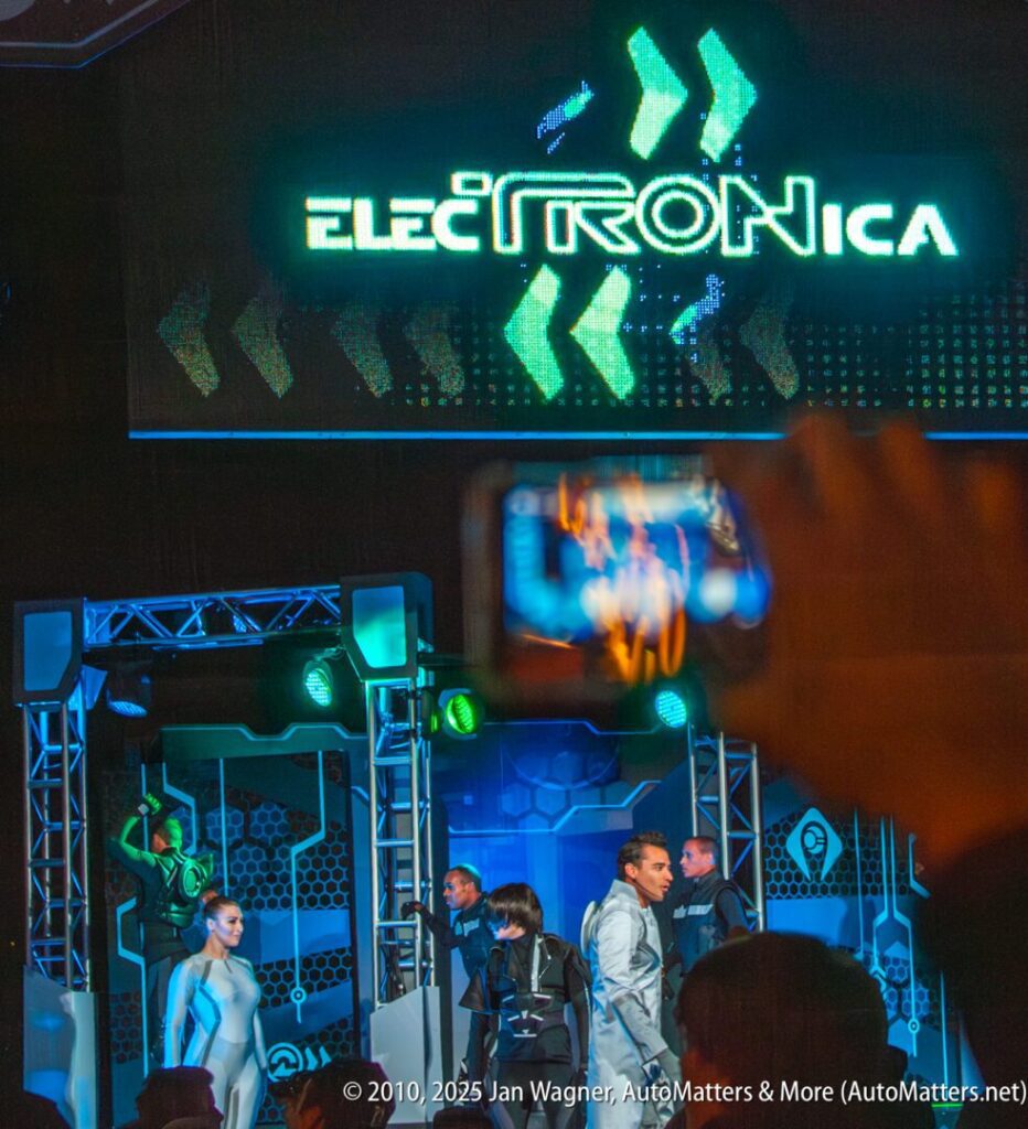 Performers in futuristic costumes stand on a stage with green lighting below a large illuminated "ElecTRONica" sign, while an audience member holds up a phone in the foreground.