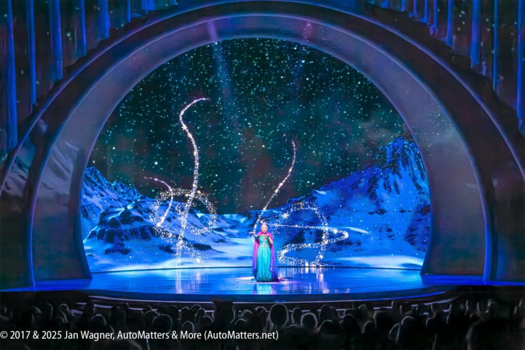 A performer in a blue dress sings on stage with a snowy, mountainous backdrop and sparkling light effects, framed by a large arch.