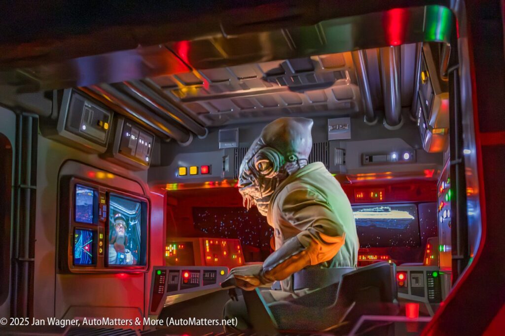 Animatronic alien character operates a control panel in a futuristic spaceship cockpit, with colorful lights and displays visible.