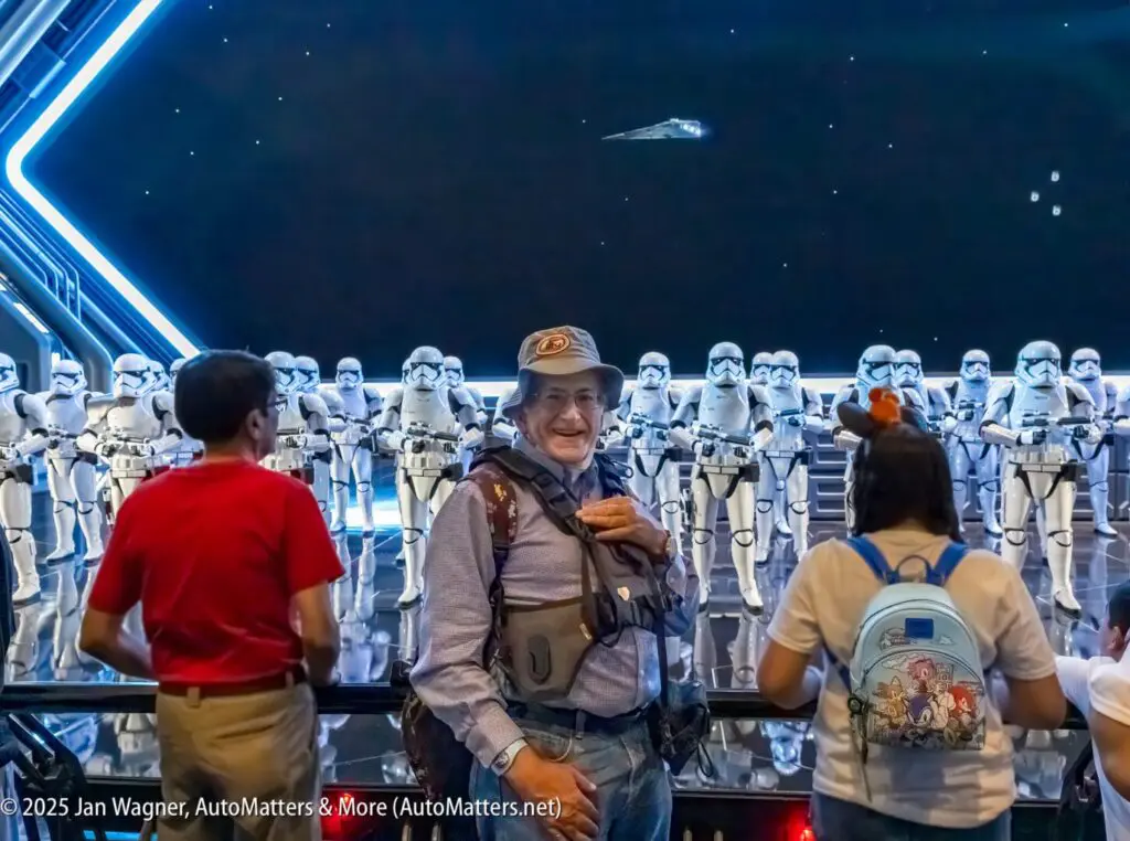 A man stands smiling in front of a group of people, with a display of Star Wars stormtroopers and a spaceship in the background.