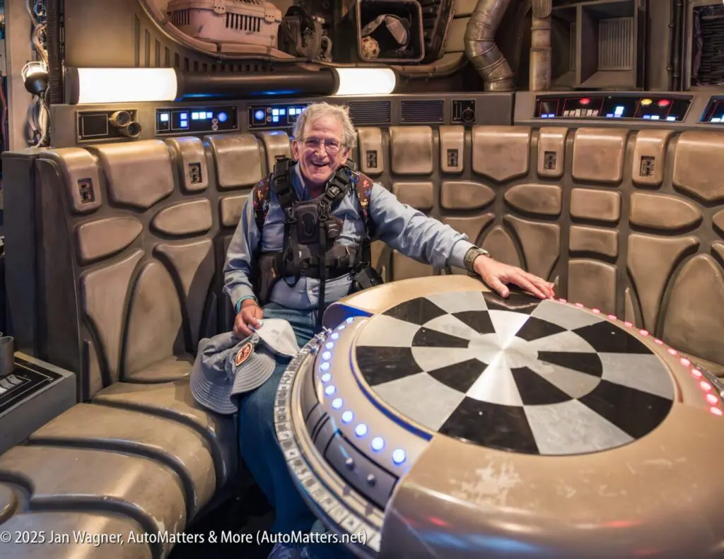 An older person smiles while sitting at a circular, black-and-white checkered game table in a sci-fi themed setting with metallic walls and controls.