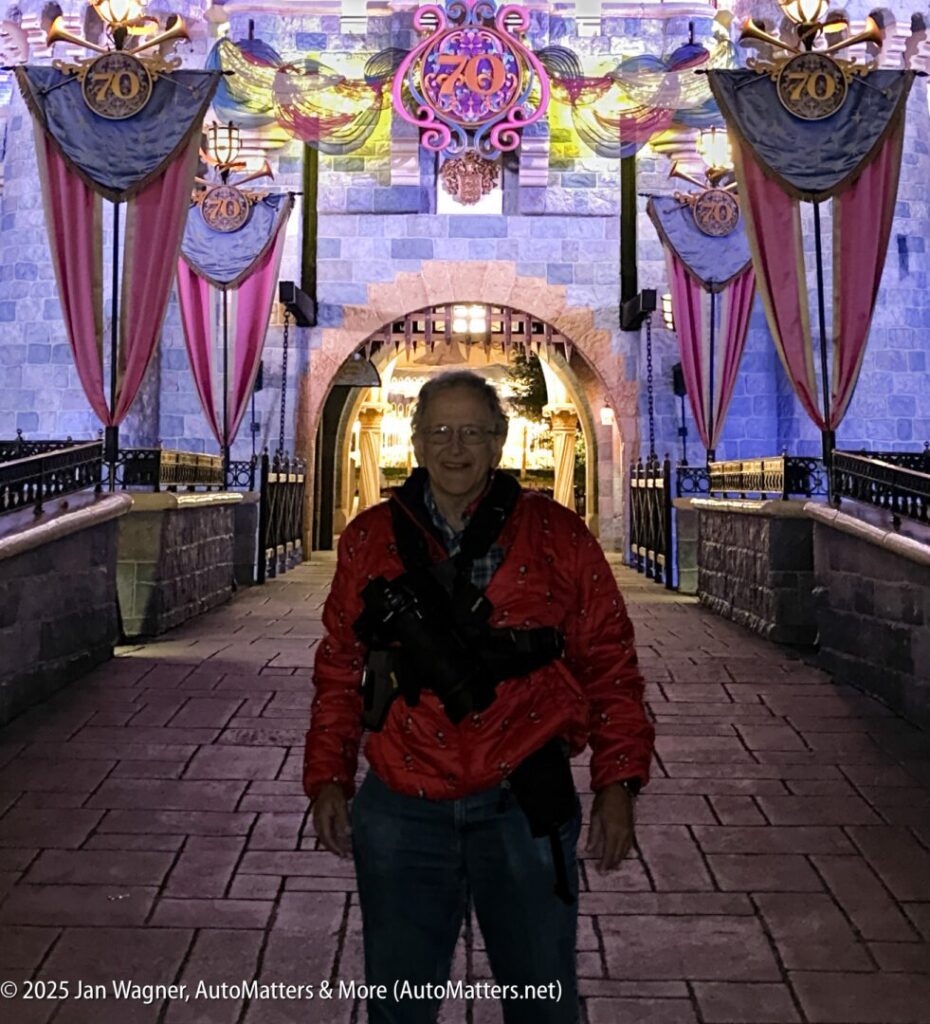 An older person in a red jacket stands smiling in front of a decorated castle entrance with "70" banners at night.