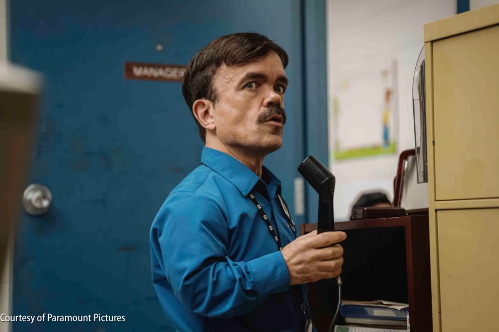 A man in a blue shirt holding a telephone handset stands near a blue door labeled "Manager" and looks to the side.