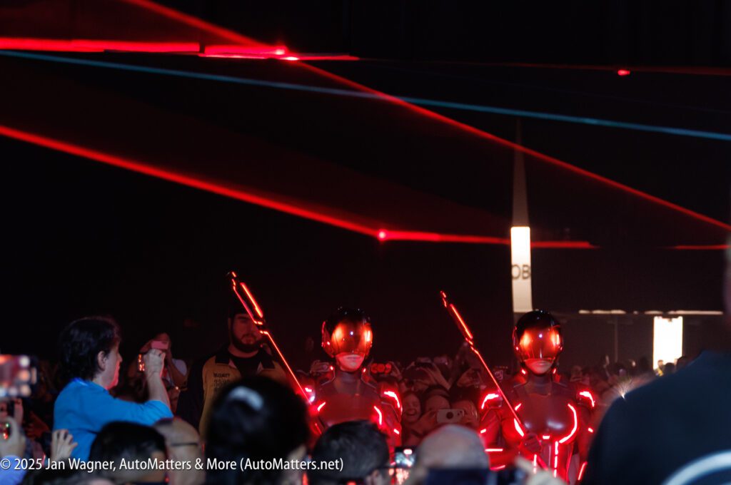 Two performers in illuminated futuristic costumes and helmets walk through a crowd under red lights at an indoor event, with spectators taking photos.
