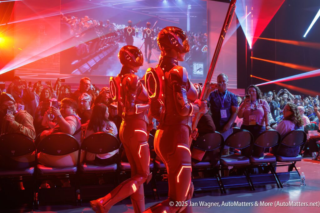 Two people in futuristic, illuminated suits walk past an audience at an indoor event with vibrant red and purple lighting.
