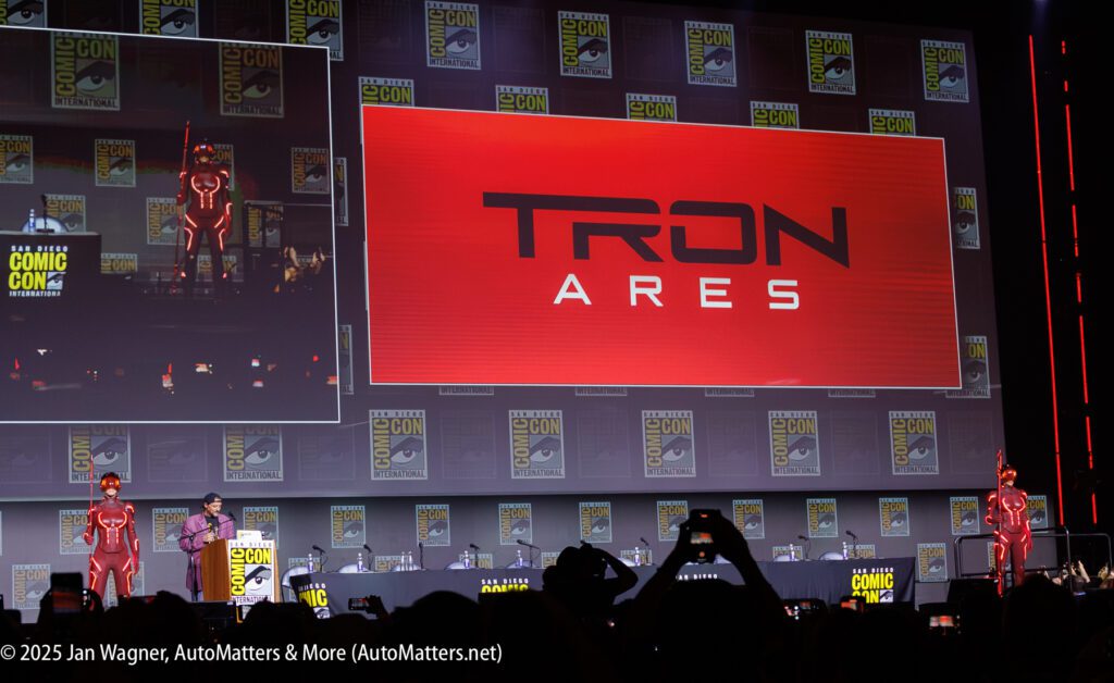 A stage at San Diego Comic-Con features presenters and two people in red TRON costumes, with a large screen displaying "TRON ARES" in the background.