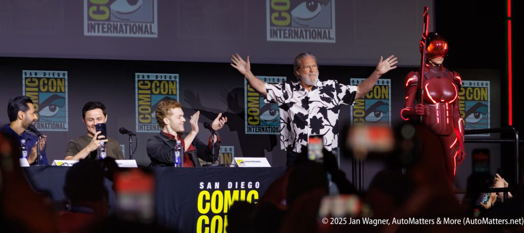 Four panelists sit at a Comic-Con table while a man with arms raised stands next to a person in a red sci-fi costume on stage. Comic-Con branding is visible in the background.