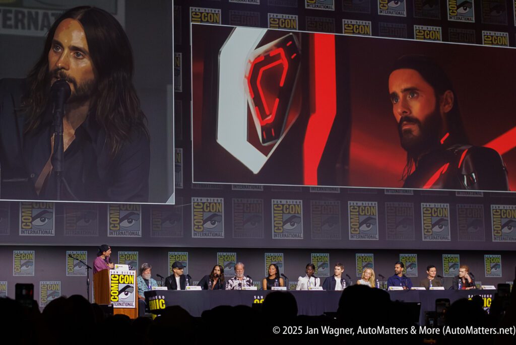 A panel of people sits at a long table on stage at Comic-Con, with a large screen showing a man with long hair speaking and a digital image of a similar man in the background.