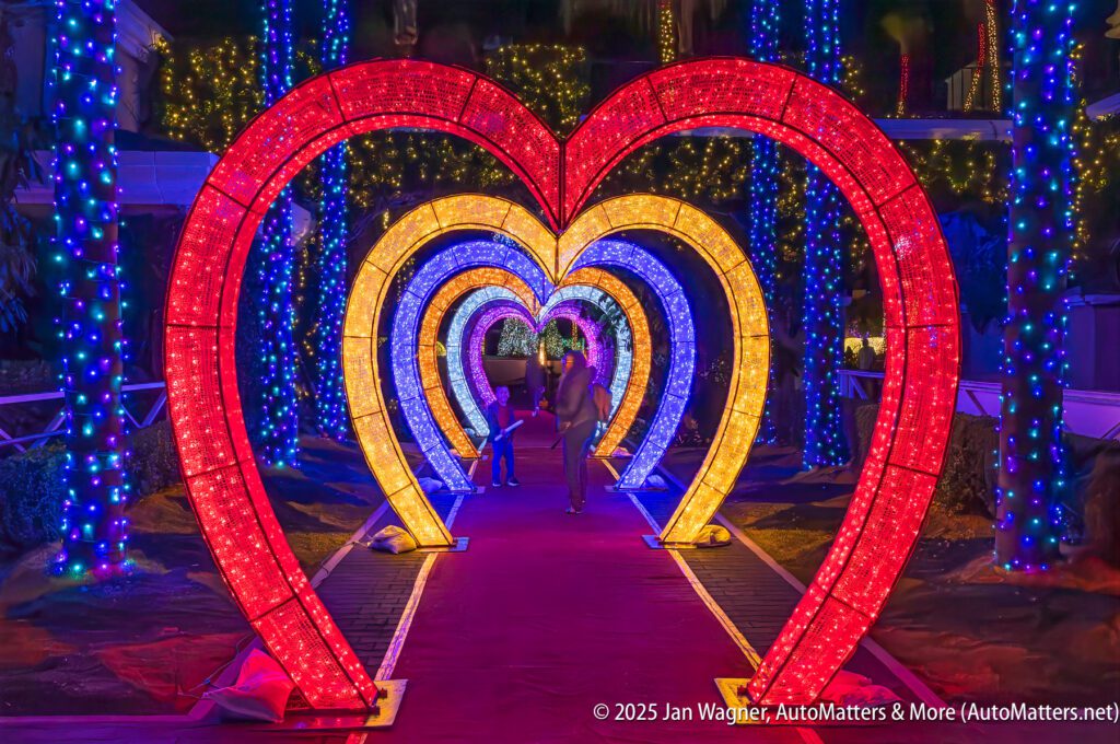 Illuminated red, yellow, and purple heart-shaped arches line a walkway decorated with string lights at night, with two people walking beneath them.