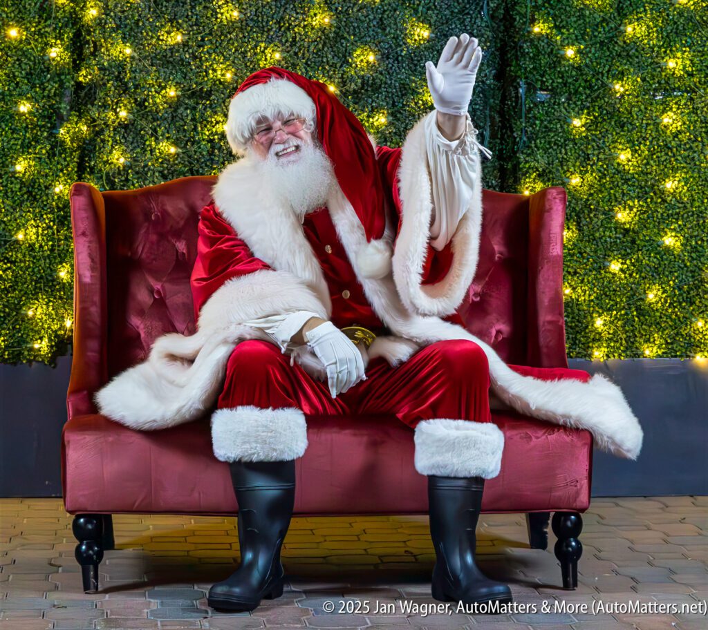Santa Claus sits on a red velvet couch, smiling and waving, with a background of green foliage and string lights.