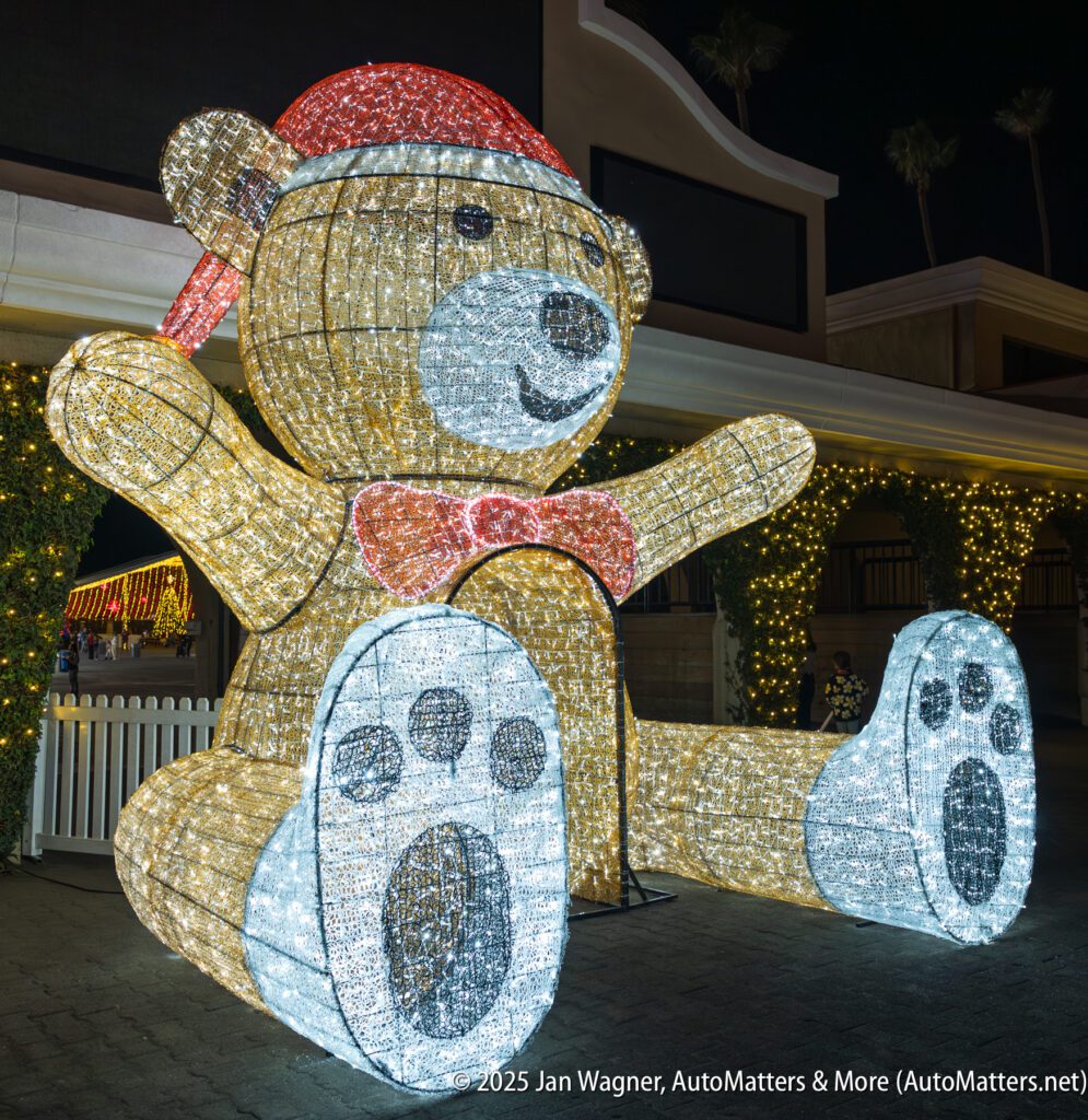 A large illuminated teddy bear decoration wearing a red Santa hat and bow tie is displayed outdoors at night.
