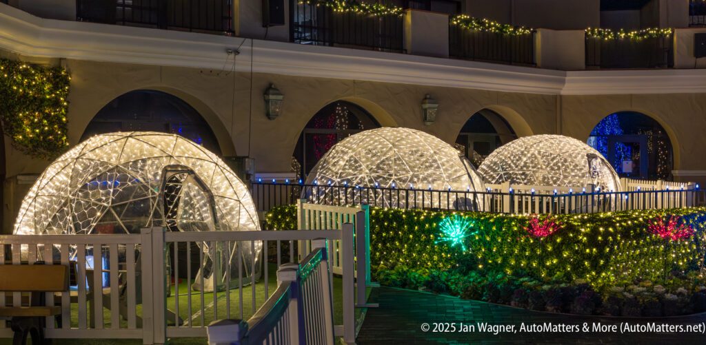 Three illuminated geodesic domes sit on a patio surrounded by white fencing and decorative lights, in front of a building with arched doorways and balconies.