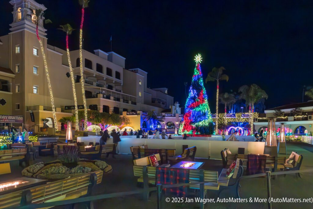 Outdoor courtyard at night decorated with holiday lights, a large Christmas tree, and seating areas with fire pits and tables.