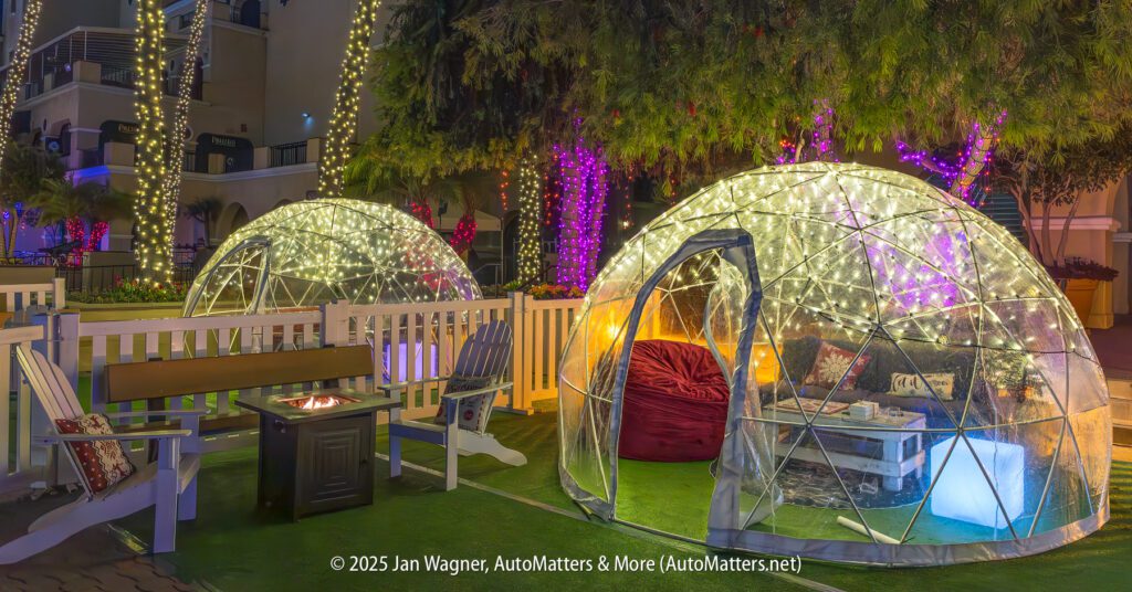 Two illuminated, transparent domes with seating inside are set up outdoors at night, surrounded by decorated trees with string lights and a nearby fire pit.