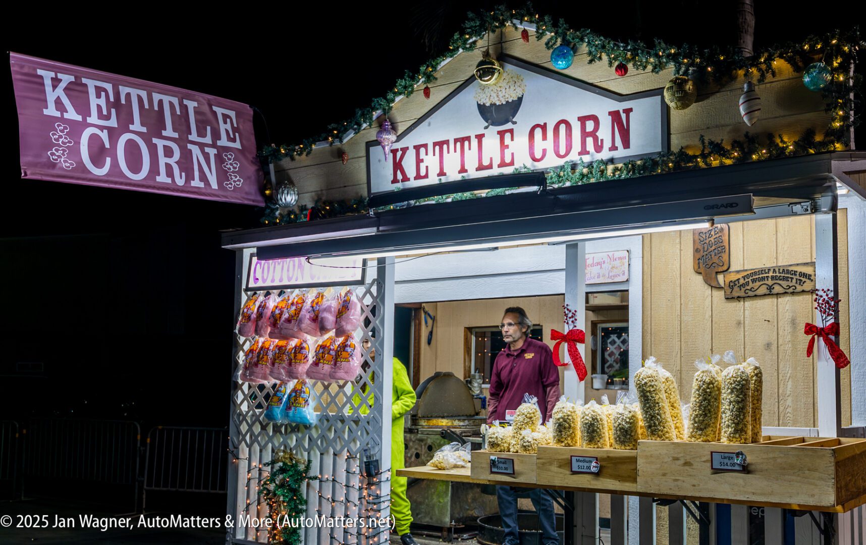 A kettle corn stand at night with bags of popcorn on display, festive decorations, and a vendor standing inside ready to serve customers.