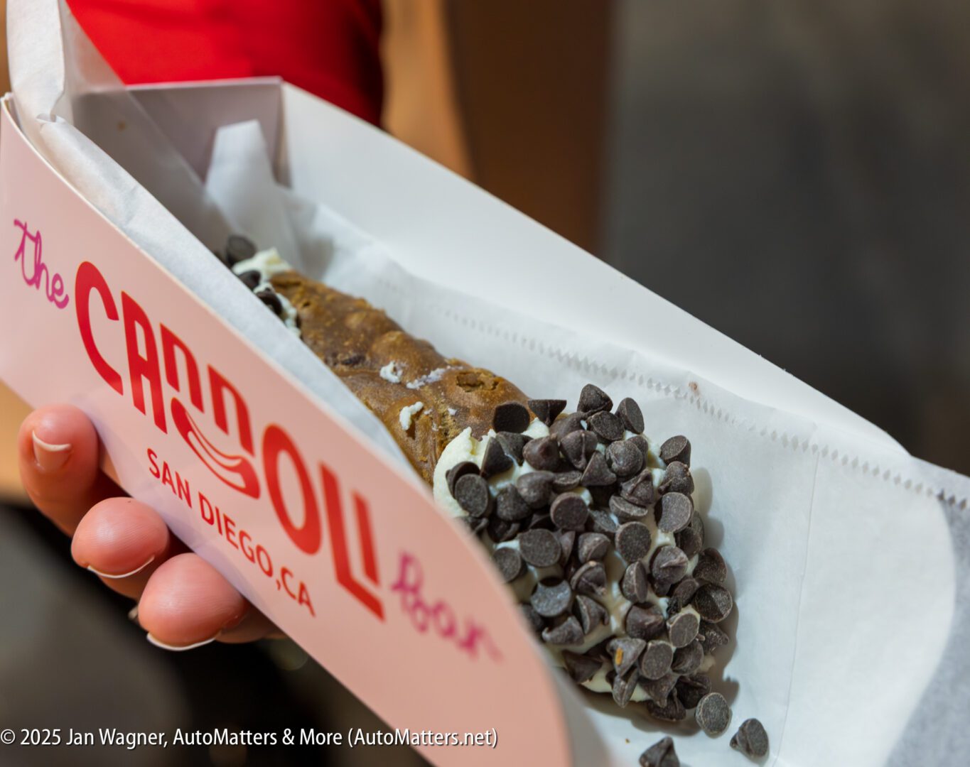 A hand holds a cannoli topped with chocolate chips in a branded box from The Cannoli Bar, San Diego.