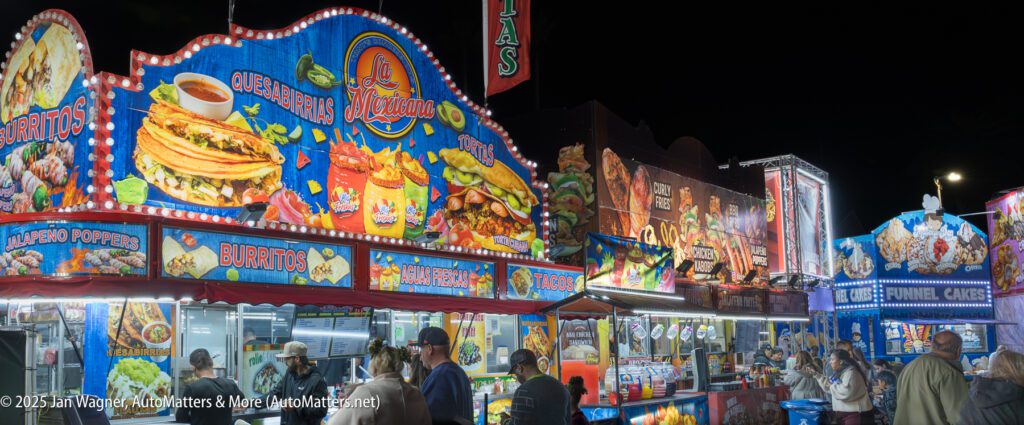 A brightly lit row of food stalls at a fair offers burritos, quesadillas, funnel cakes, and drinks at night. Crowds of people order and wait in front of the booths.