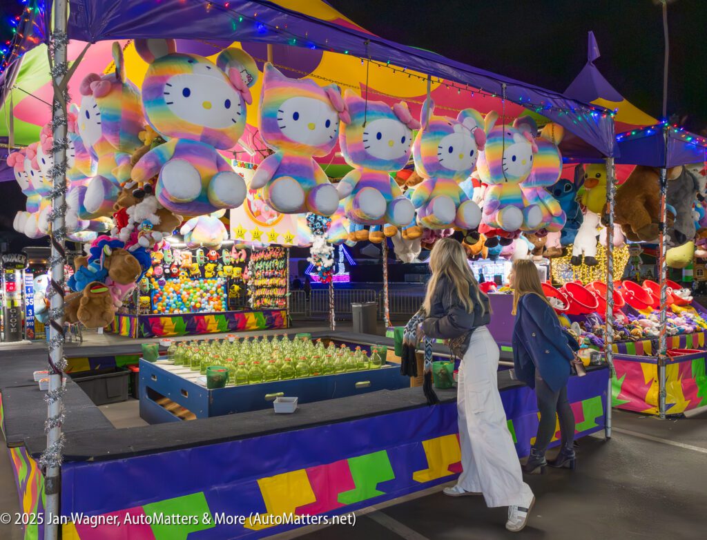 Two women stand at a brightly lit carnival game booth at night, with large rainbow Hello Kitty plush toys hanging above as prizes.