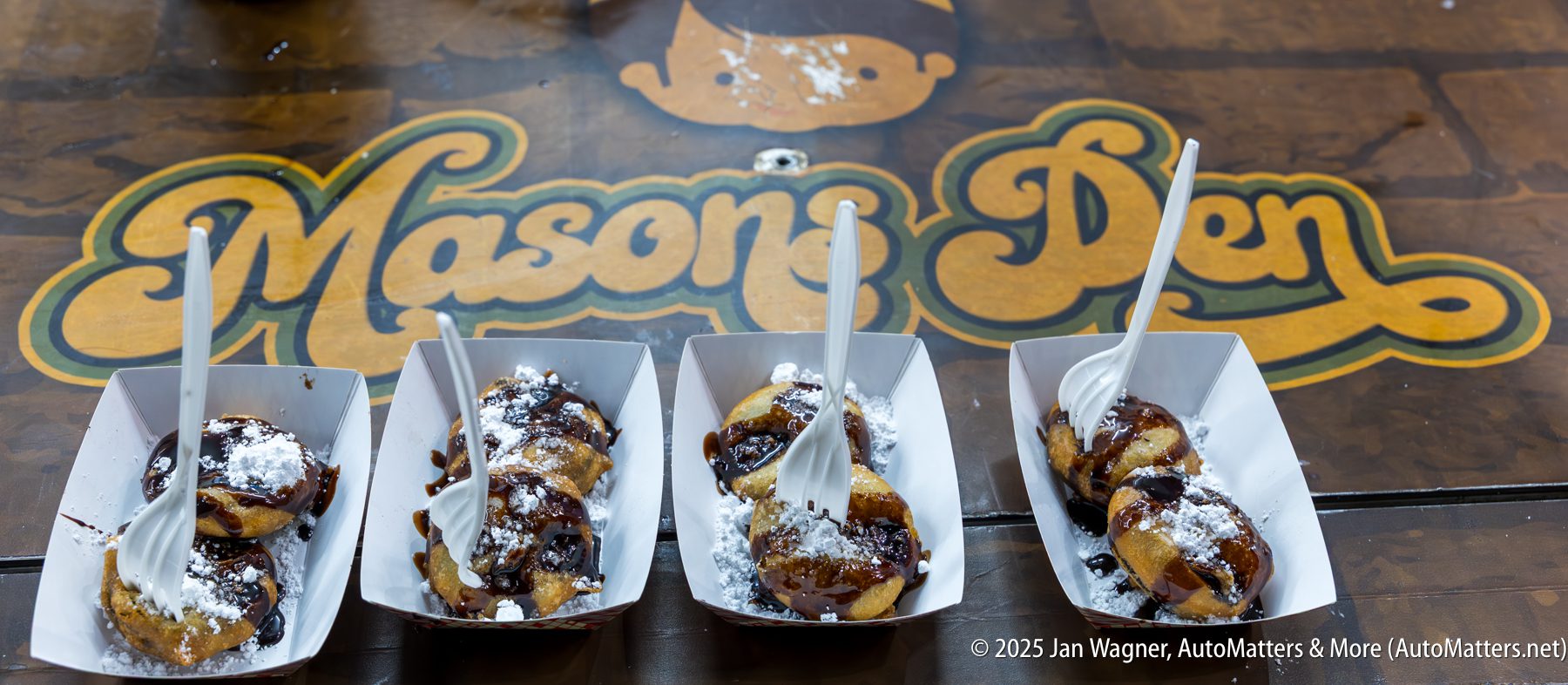 Four trays of powdered sugar and chocolate-drizzled desserts with plastic forks are placed on a table with a "Mason's Den" logo in the background.