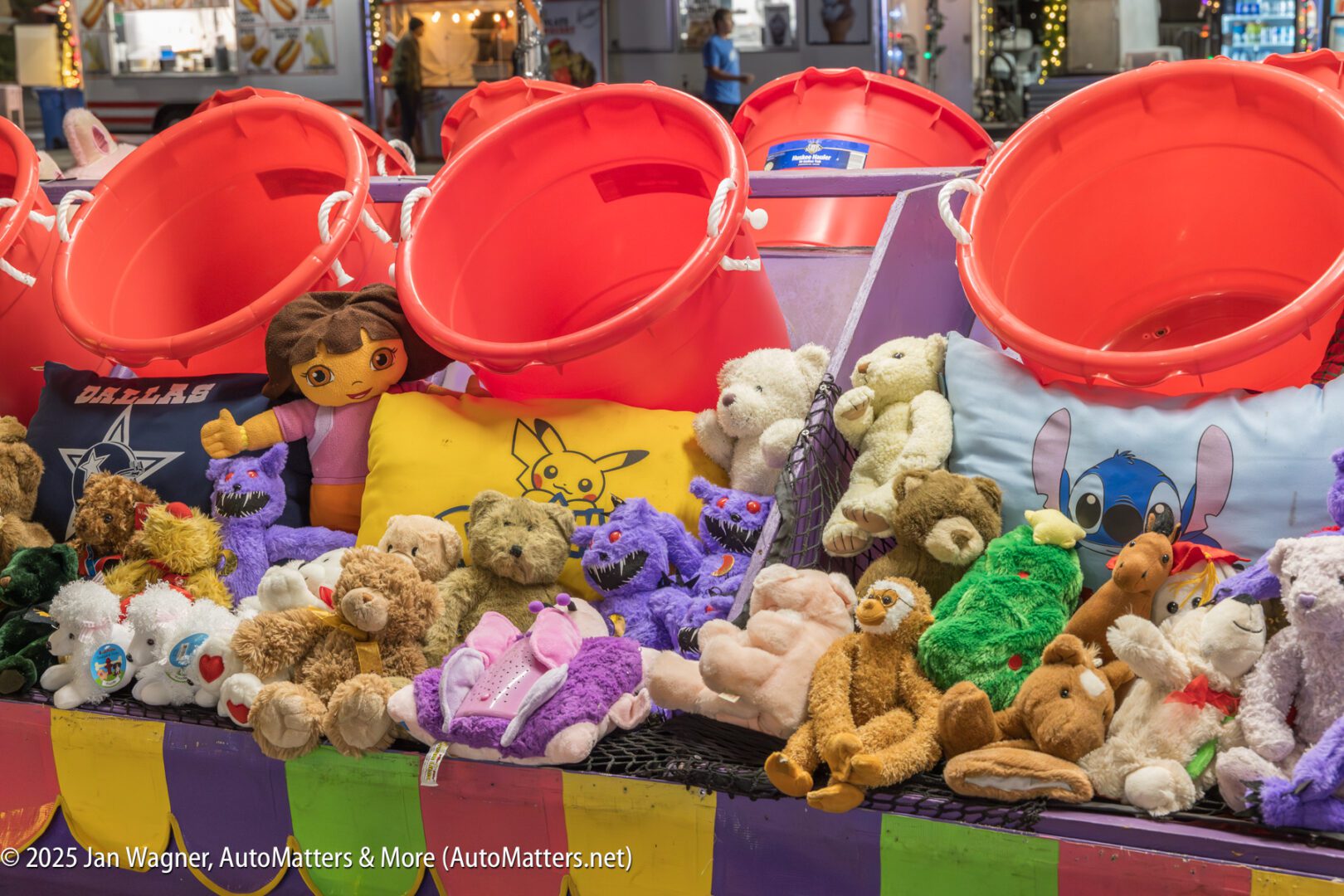 Carnival game booth with large red buckets, colorful plush toys including animals and character pillows as prizes, and festive lights in the background.