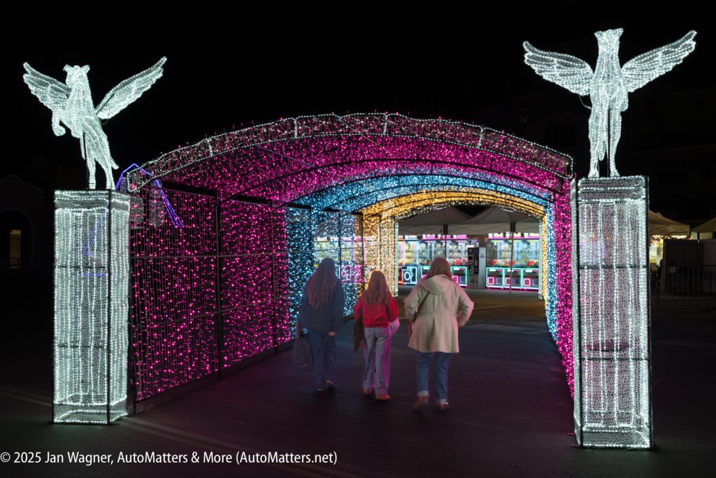 Three people walk under a tunnel of colorful, illuminated lights with winged horse statues on pedestals at the entrance, at night.