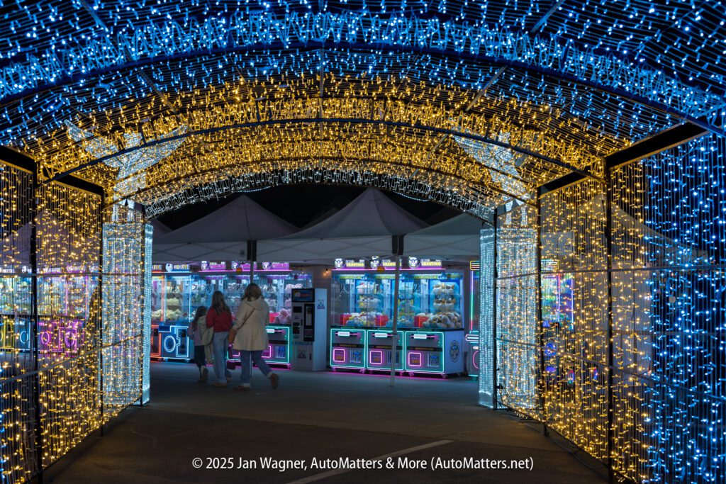 A tunnel of blue and yellow holiday lights leads to an area with claw machine arcade games and a few people walking at night.