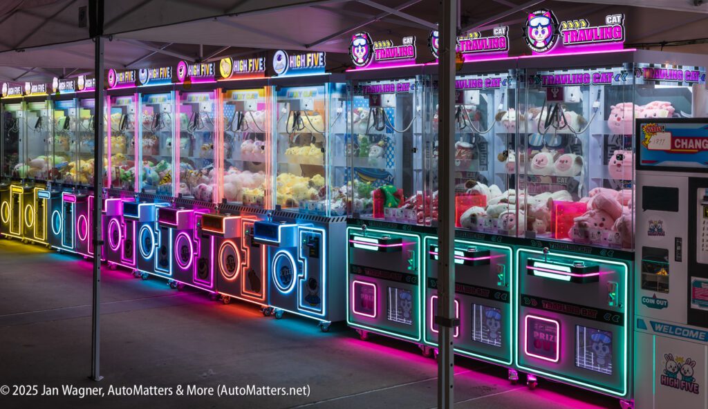 A row of brightly lit claw machines filled with plush toys is set up outdoors under a tent at night.