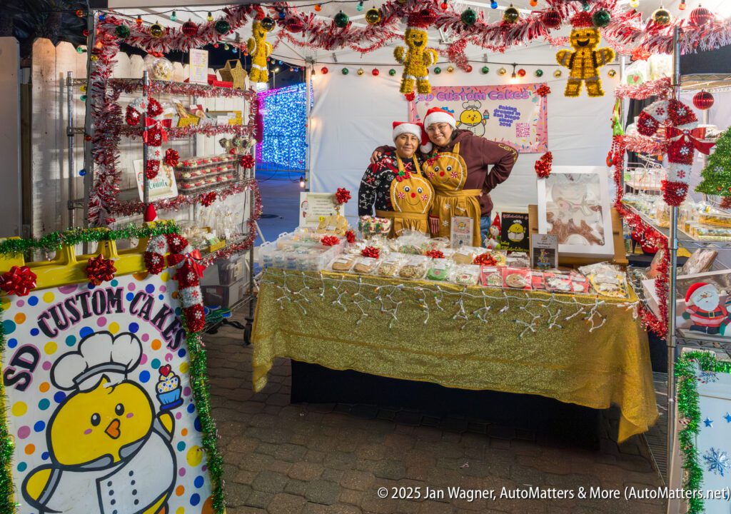 Two people stand behind a decorated table at a festive outdoor market booth selling cookies and cakes, featuring yellow chick and gingerbread man decorations.