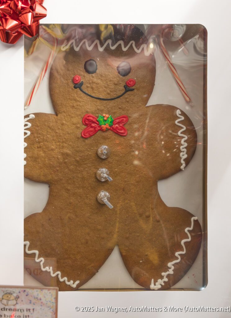 A large gingerbread cookie decorated with white icing, red buttons, a green bowtie, and three silver Hershey's Kisses, displayed in a box with a red bow on the corner.