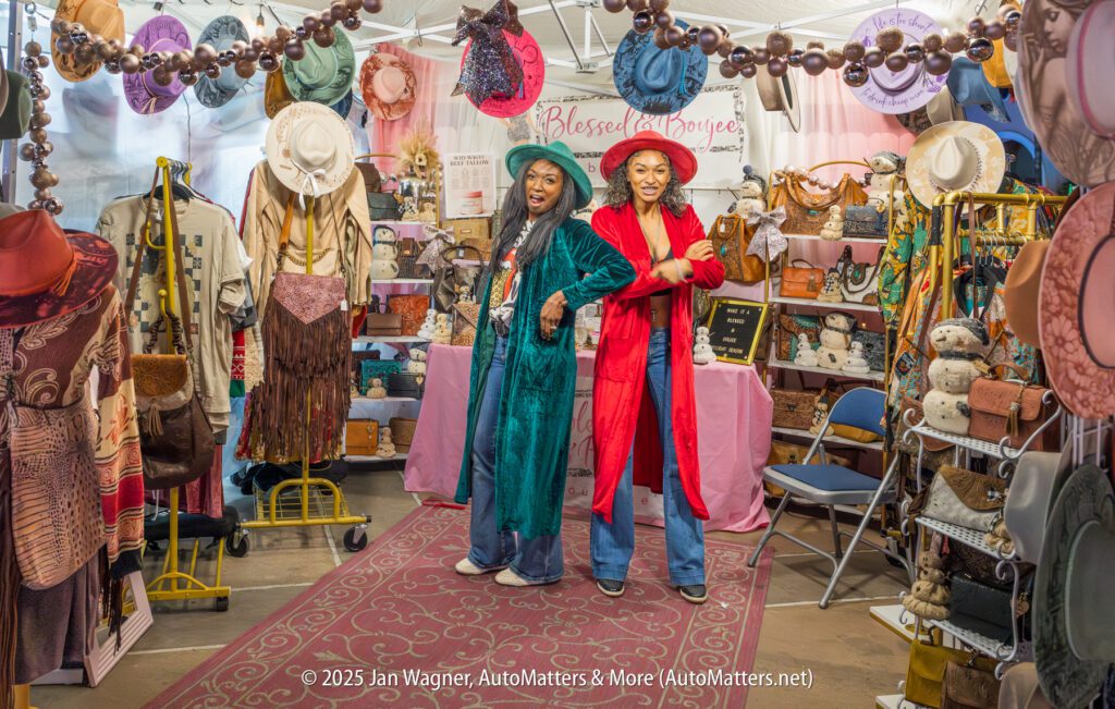 Two women in colorful coats and hats stand and pose inside a busy boutique filled with hats, bags, and various accessories on display.