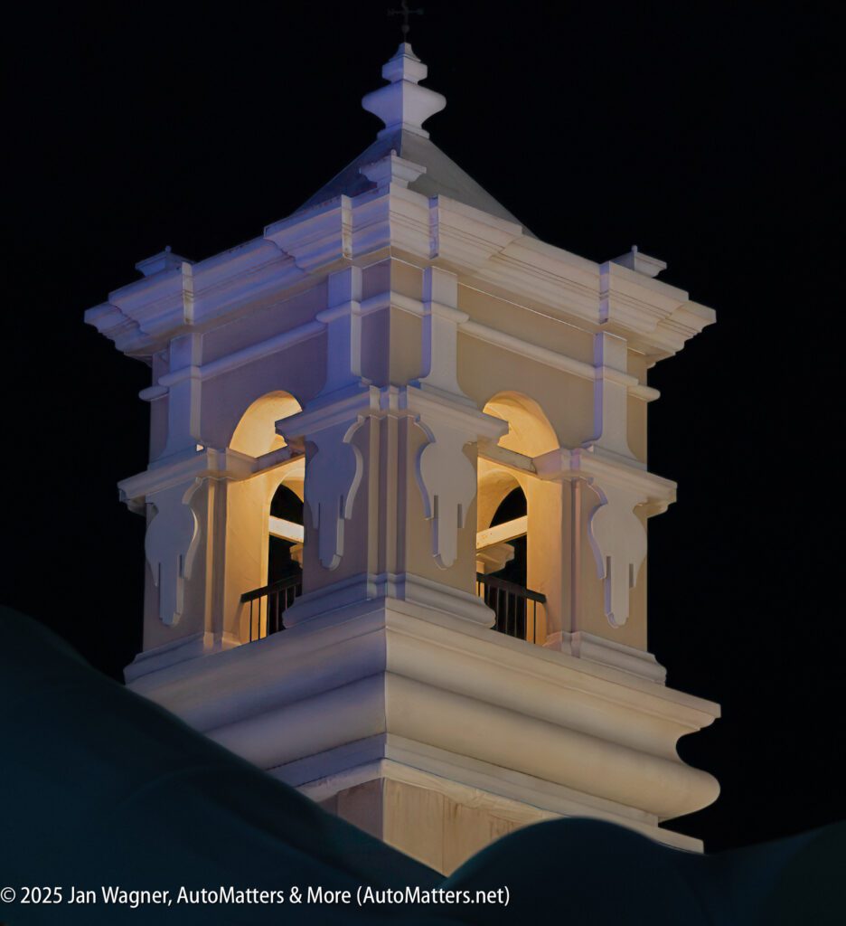 A white bell tower is illuminated against a dark night sky, with part of a roof visible in the foreground.