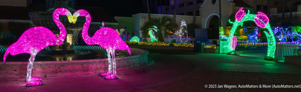 Two illuminated pink flamingo decorations stand near a fountain, surrounded by colorful holiday lights and displays at night.