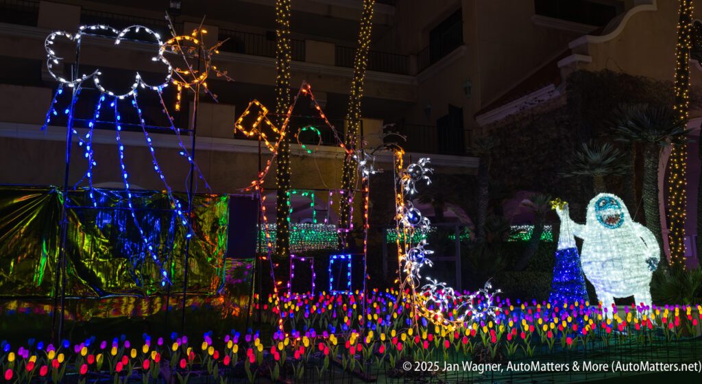 A festive outdoor display with colorful LED lights forms a house, a cloud with raindrops, tulips, and a large yeti figure, set against a building at night.