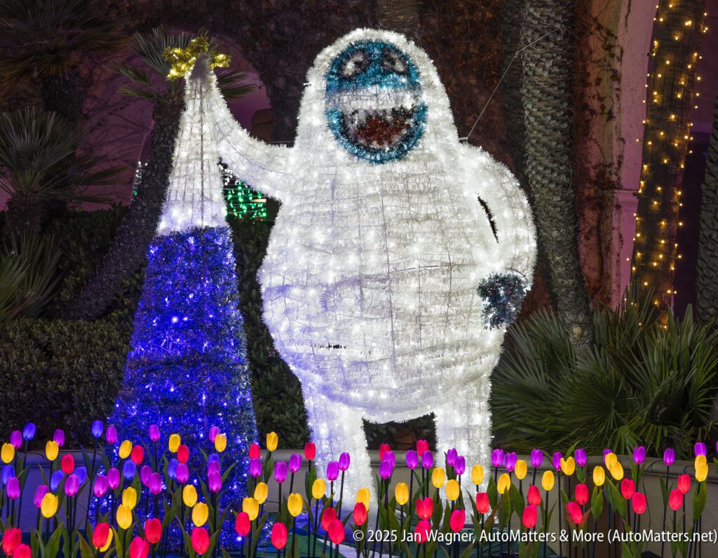 A large illuminated abominable snowman decoration stands next to a blue and white tree, surrounded by artificial colorful tulips at night.
