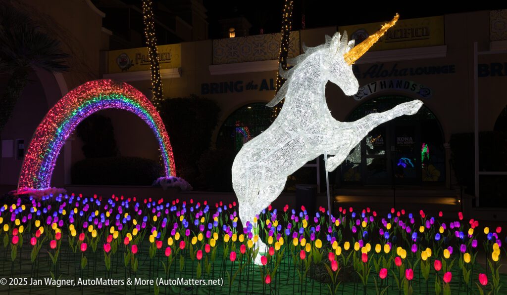 A large illuminated unicorn sculpture stands among colorful artificial tulips, with a bright rainbow light display in the background at night.