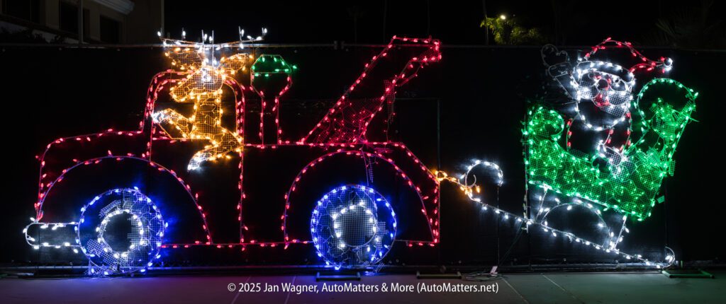 A holiday light display shows a reindeer driving a tow truck pulling Santa Claus in a sleigh, illuminated in various colors at night.