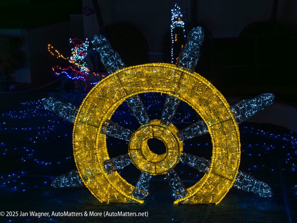Large illuminated ship wheel decoration with yellow and blue lights, surrounded by other light displays, set against a dark background.