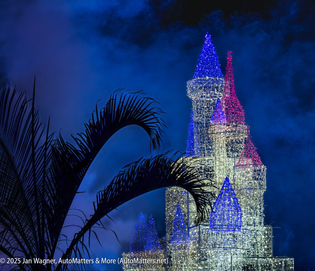 A wireframe castle sculpture illuminated in white, pink, and blue lights stands behind a silhouetted palm frond against a dark, misty sky.
