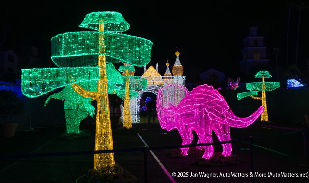 A nighttime display of illuminated sculptures features green trees, a pink dinosaur, and a castle, all made from colorful lights.