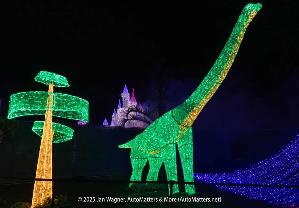 A large green dinosaur and a tree are outlined in colorful lights at night, with a castle shape illuminated in the background.