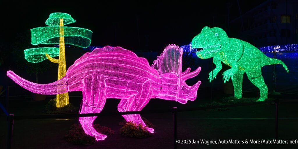 Two large dinosaur figures, a triceratops and a T. rex, made of colorful lights are displayed outdoors at night, with a stylized light-up tree in the background.